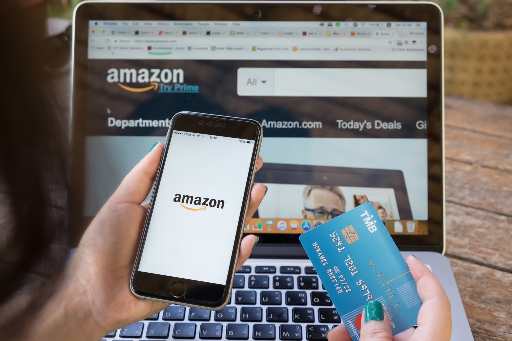 A person holding an iPhone displaying the Amazon logo and a TMB credit card, with an Amazon webpage open on a laptop in the background.