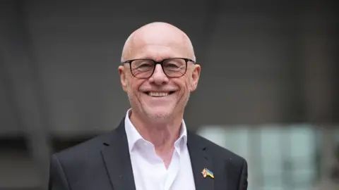PA Businessman John Caudwell leaving BBC Broadcasting House in London - he is wearing a black dress jacket with a Union Flag and Ukraine flag pin on his lapel - he has a bald head, a broad smile and is wearing dark rimmed glasses, with his white shirt unbuttoned and with no tie 