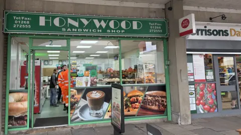 Elliot Deady/BBC The exterior of a sandwich shop. Above the door is a green sign with the business name - Honywood Sandwich Shop - in white lettering. About five people can be seen inside the shop.