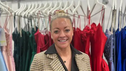 Lauren Carter/BBC A smiling woman - Kristy Chamberlain - looking straight at the camera. She is wearing a patterned blazer over a black dress and is standing in front of a rail of prom dresses.
