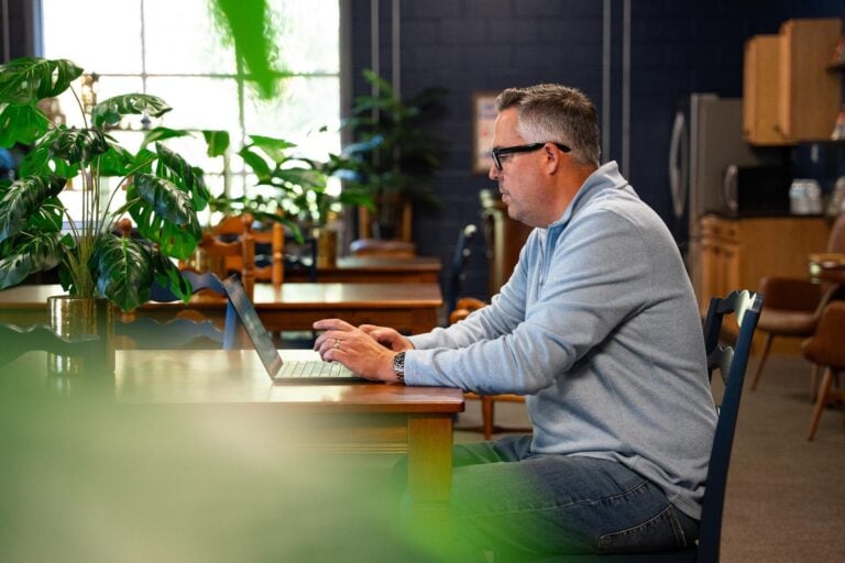 A white man sits behind a laptop on a wooden table, viewed from the side in a well-lit room with many plants.