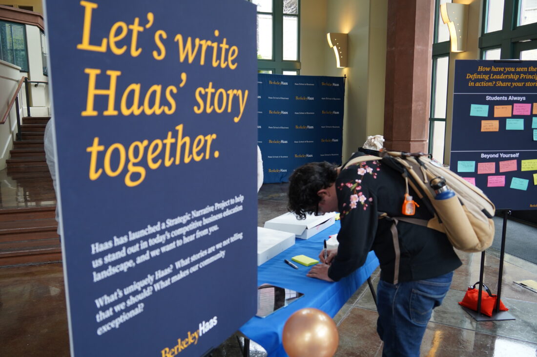 a student wearing a backpack leans over a table to write a on a post it next to a sign that says: Let's write Haas' story together.
