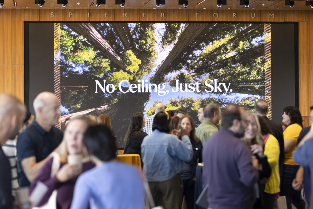 A crowd of people in a room with a large screen that reads: No Ceiling, Just Sky and shows a photo of redwood trees.
