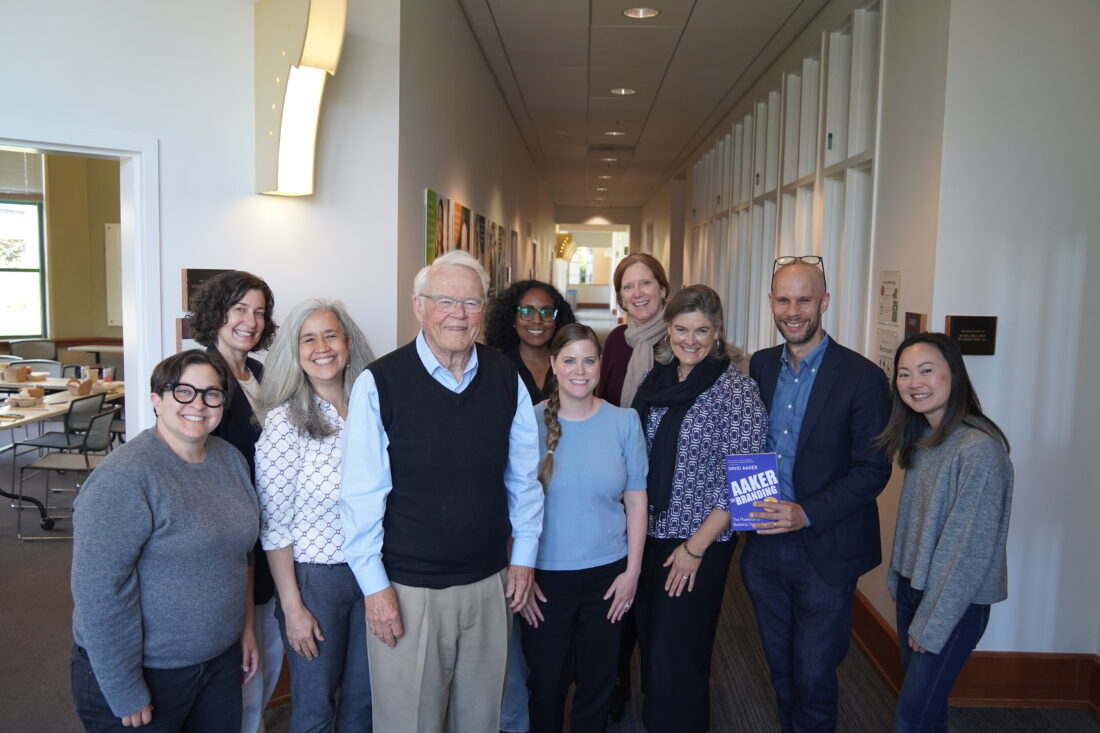 A group of smiling people stand in a hallway, flanking a smiling man with gray hair and a navy blue sweater vest.