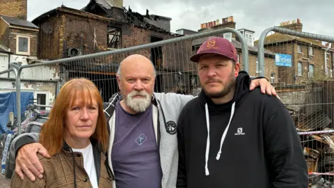 BBC/Wendy Hurrell Three people, a woman on the left with red hair, a man with a grey beard in the centre and younger man wearing a cap look sadly into the camera with the wreak of a burned building in the background
