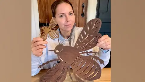 Rustic Warehouse Norfolk Louise Banham holding up two rusty coloured bees, with wings outspread, in front of her. One is large and measures 51cm across and she is holding it in her left hand. In her right hand, she holds up a much smaller bee. She is sitting at a table, wearing a light grey sweatshirt. Her dark hair is pulled back from her face. 