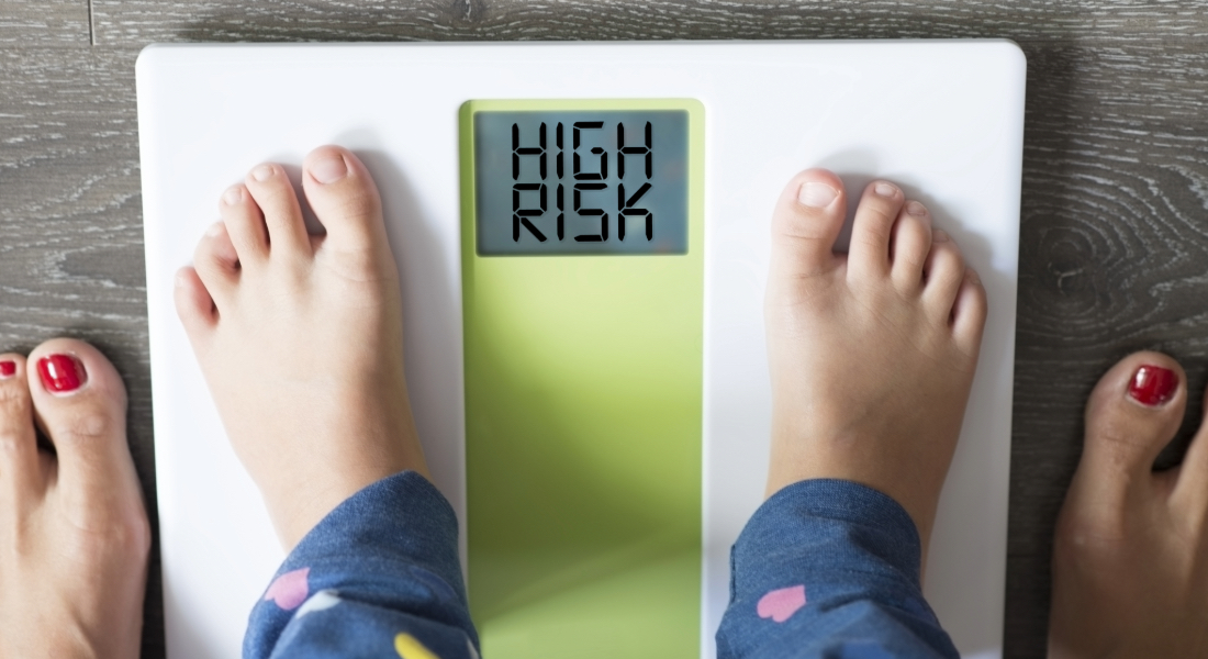 A photo of a child's feet on a weight scale with a woman's feet behind them and the words "high risk" on the scale display.
