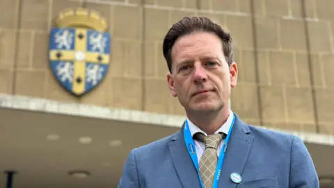 BBC Durham County Council leader Andrew Husband outside County Hall. He is wearing a light blue suit jacket, a white shirt and a gold tie. His brown hair is swept back.