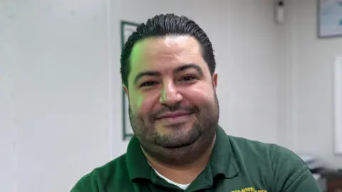 Elliot Deady/BBC A man - Murat Mutlu - looking straight at the camera with a happy expression. He is standing in a sandwich shop and is wearing a green polo shirt with the name of the shop - Honywood Sandwich Shop - embroidered in yellow.