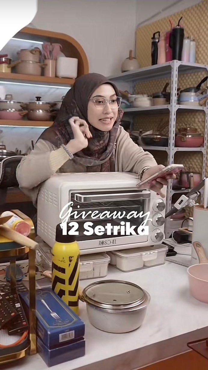 A woman wearing glasses and a hijab enthusiastically gestures while presenting a giveaway for 12 irons, standing at a kitchen table surrounded by pots, cooking utensils, and kitchen items.