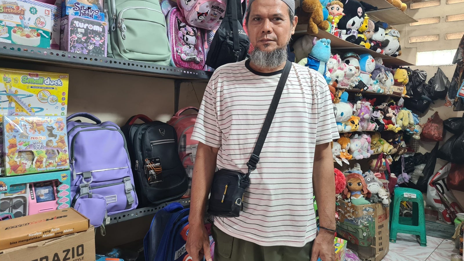 A man with a beard wearing a striped shirt and a cap stands in a colorful shop filled with backpacks and plush toys, with shelves displaying various children's toys behind him.