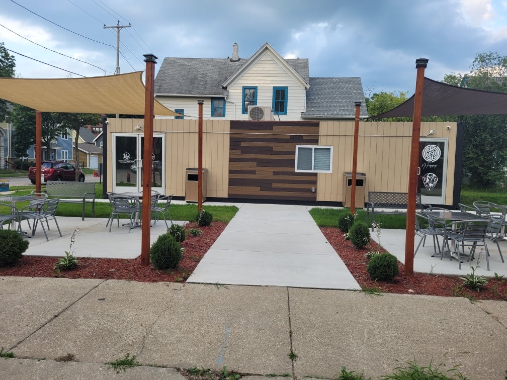 Outdoor seating area with metal tables and chairs, shaded by fabric canopies, facing a modern building with wood panel accents and decorative signs on the doors.