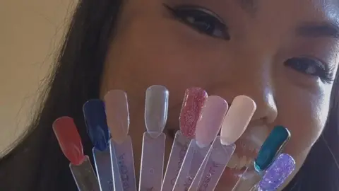 Kintsugi Beauty A close up shot of a woman smiling. She has black hair and is holding up fake nails on plastic sheets which she has painted in shades ranging from red to blue.