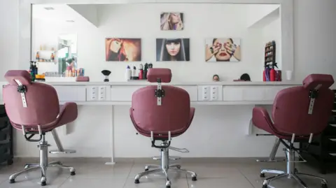 Guilherme Petri A stock image of an interior of a white beauty salon with a tiled floor. Three wine coloured swivel chairs are facing a mirror. Four paintings of women's faces can be seen in the reflection. Various hairspray bottles and makeup equipment are on the table below the mirror.