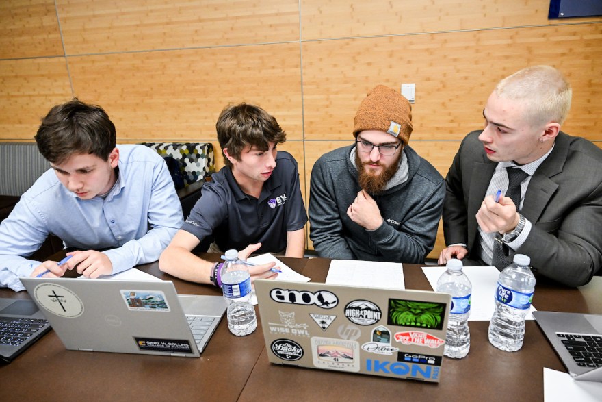 Students sit at a table talking and looking at their computers. 