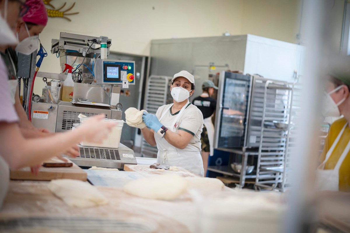 Bakery workers, wearing protective masks, prepare dough in an industrial kitchen. One worker in the center holds a piece of dough, while others are focused on different tasks.
