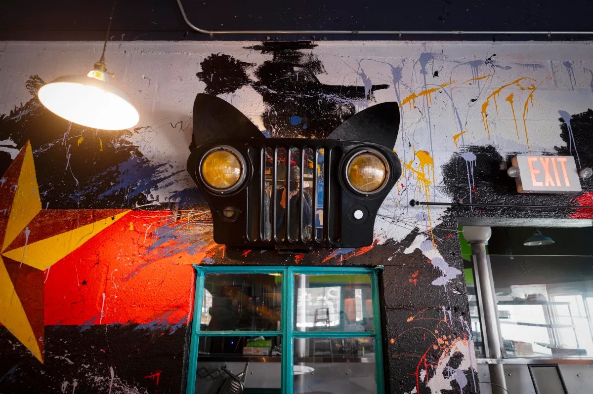 A wall decorated with a car grille and headlights arranged to resemble a cat’s face, surrounded by bright, abstract graffiti art.