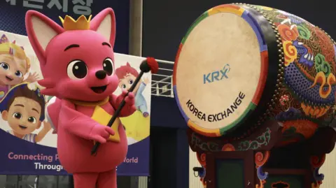Pinkfong A photo of a Pinkfong mascot hitting a drum on stage to mark the launch of the company's shares on the stock market. The words "Korea Exchange" and its logo are printed on the multi-coloured drum.