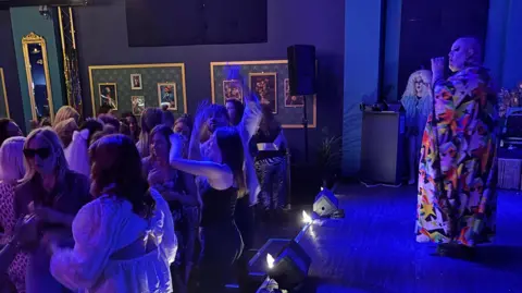 Dragonfly Lounge A drag performer is singing on stage in a colourful gown, with people dancing.  The room is lit in blue.