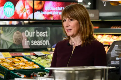 PA Media Rachel Reeves stands in front of the fruit aisle in a Tesco supermarket wearing a red zip-up top