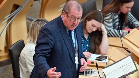 Getty Images A man with thinning grey hair and glasses, wearing a blue suit, speaks in the parliament chamber. 