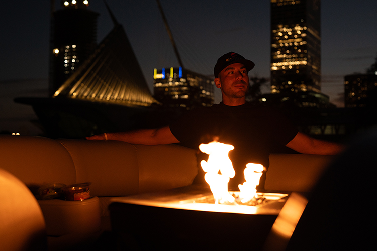 A man sits at night in front of a fire pit with a flame in it 