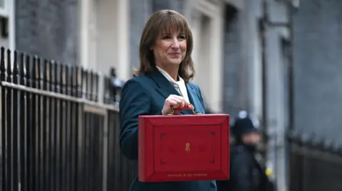 A woman with shoulder-length hair standing outside No10 Downing Street, next to black railings. She is wearing a navy suit and holding a red briefcase whilst smiling. 