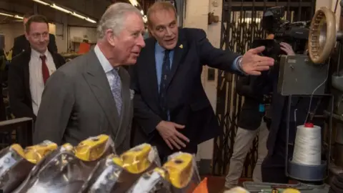Getty Images Inside a shoe factory King Charles is wearing a brown suit with a lilac-coloured tie. He is standing next to another man in a navy suit who is pointing to a manufacturing machine. Several shoes in the process of being made are on display