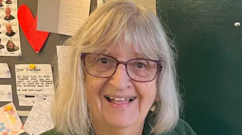 Jane Calcott A woman standing in front of a notice board smiling. She has light shoulder-length hair and is wearing glasses. Behind her are notes pinned to a board 