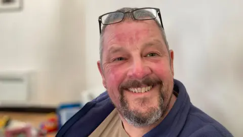 Mike Carson wearing a navy jacket with a beige top. He has a moustache and beard and is smiling. His glasses are resting on top of his head. 