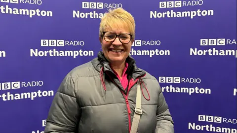 A woman wearing a silver coat with a pink top and standing in front of a purple backdrop which reads BBC Radio Northampton. She has short blonde hair, is wearing glasses and is smiling. 