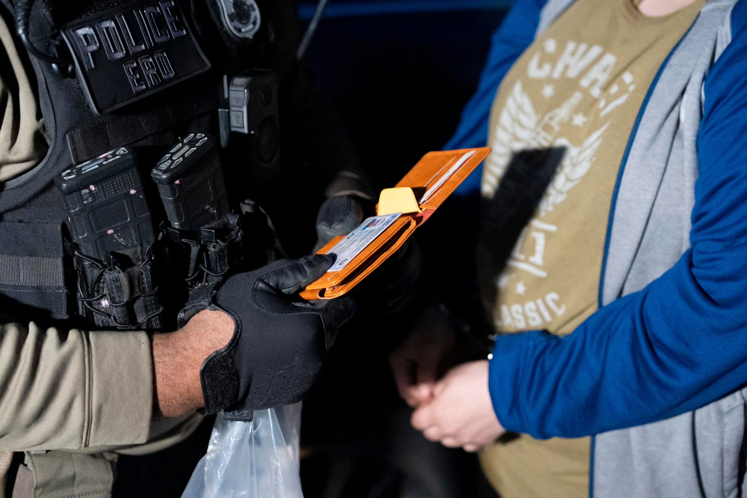 A law enforcement officer in tactical gear examines an ID card from a wallet while another person stands nearby with hands clasped.