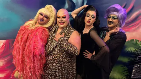 Dragonfly Lounge Four drag artists posing for the camera in front of a mural. From left to right: One is wearing pink feathers and a blonde wig; one in leopard print; a dark haired drag artist in a black laced dress and pearls; and on the end in black, is one with a purple wig and moustache.