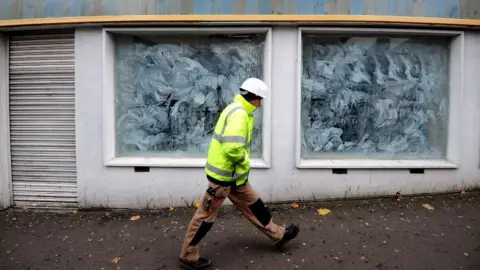 PA Media A man in a high-vis jacket, white hard hat and brown trousers walks past a close shop front, with shutters down on the door and windows covered in white paint. 