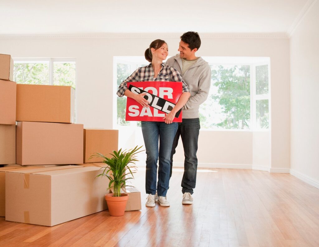 Couple holding a for sale sign in living room with moving boxes.