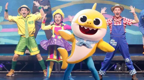 Getty Images A performer in a yellow Baby Shark costume dances beside others dressed in colourful outfits onstage during "Pinkfong Baby Shark Live!" show in New York