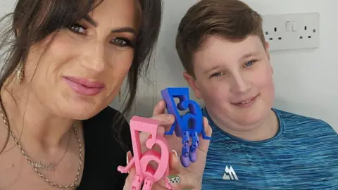 Handout Karlla, a woman with dark hair, andAlfie, a pre-teen boy, sit side-by-side. They are each holding up small plastic statues of the number '67' with little arms and legs attached. The numbers appear in reverse, as in a mirror image, as they have taken a selfie.