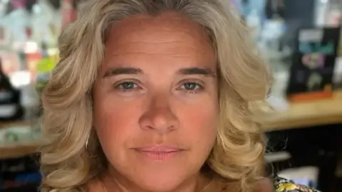 Miranda Richardson Miranda Richardson smiles at the camera. She is standing behind a bar with bottles of alcohol behind her. She has curly blonde hair and is wearing hoop earrings. 