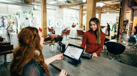 Blake Wisz A stock image showing the interior of a bright, modern hair salon. In the foreground, two women are interacting at a counter — one stands behind it, smiling, while the other faces her from the customer side, holding what looks like a payment terminal. The counter has a computer screen and some small items on display. Behind them, the salon is open and well-lit with large mirrors, chairs, and people working or chatting. The space features wooden beams, exposed ceilings, and large windows letting in natural light.