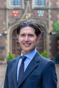 Christopher Marquis stands outdoors in front of a historic building with crests on the wall, wearing a suit and tie, smiling.