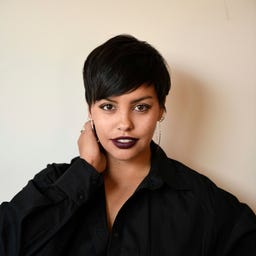 Alex Temblador wears a black shirt and stands in front of a beige wall with her hand on her neck