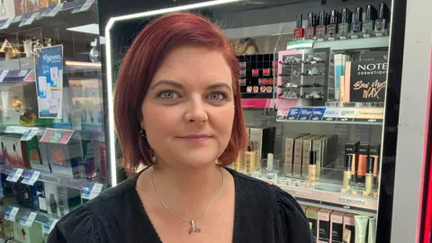 A woman with short red hair stands in front of makeup and fragrance shelves inside a shop. She is wearing a black top and a necklace.