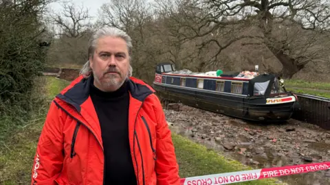 A man with grey hair and facial hair with a red coat standing next to a canal which has no water. A narrowboat is on the bed of the canal and it is half hanging over the edge of a large hole