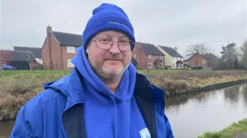 A man with greying facial hair and glasses is standing in front of a canal. He is wearing a blue beanie hat, hoodie and jacket. The beanie has white writing that reads floating holidays 