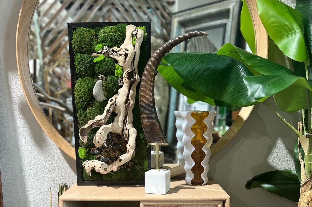 A cabinet in front of a mirror and next to a houseplant with large leaves. There are a sculpture, a vase and a piece of moss art sitting on top of the cabinet.