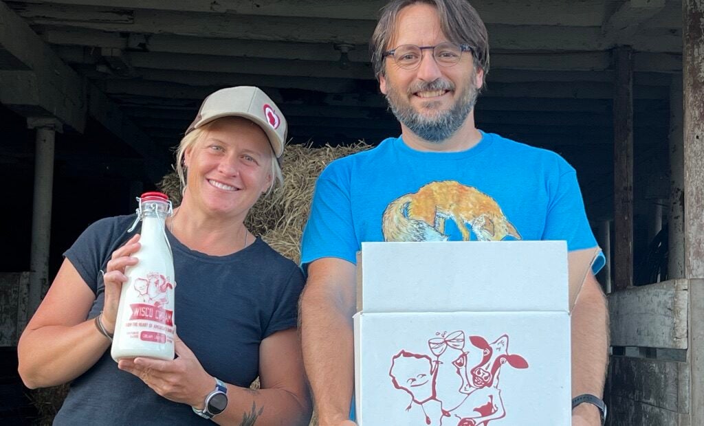 Two people stand in a barn holding Wisco Cream dairy products; one holds a glass milk bottle, the other holds a box labeled Wisco Cream, From the Heart of Americas Dairyland.