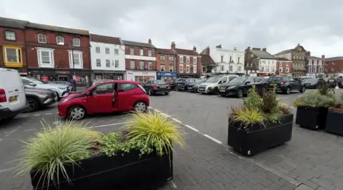 A view of a large market place, laid with grey block paving stones, with parking in the centre and black planters, topped with green shrubs, at the front  of the photo. Cars are parked behind them. The square is surrounded by buildings, some Georgian-style, some more modern retail units.