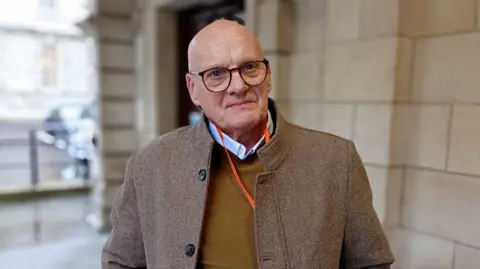 Ian McLaughlin is wearing glasses in a brown wool coat and yellow jumper. He's standing in front of a cream stone building.