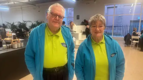 Godfrey Brock-Gadd and Hazel McLean both the grey hair. They are both wearing green T-shirts and turquoise fleeces. There is a cafe in the background. 