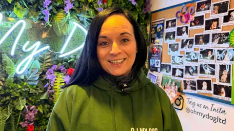 BBC / Jasmine Lowe A woman smiling at a camera as she stands in a brightly decorated shop. She has black hair and is wearing a green hoodie. She is standing in front of a wall of fake plants, with a neon sign to her left. To her right are lots of Polaroid-style photos of dogs which are pinned up on the wall.  